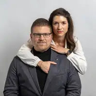 Ein Pärchen in schicker Kleidung. Die Frau steht hinter ihrem Mann und umarmt ihn